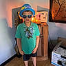 Kingston is registered to the contest to win money with this photo: child, boy, hat, sunglasses, flower_lei, turquoise_shirt, shorts, sandals, indoor, wooden_floor, cabinet, decor, lamp, shadow, picture_frame, box, plant, wall_art, portrait, casual_clothing