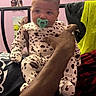 baby, pacifier, onesie, hand, person, indoor, chair, clothing, purple_wall, poster, bed, blanket, adult, sitting, infant, cute, furniture, portrait, people, casual