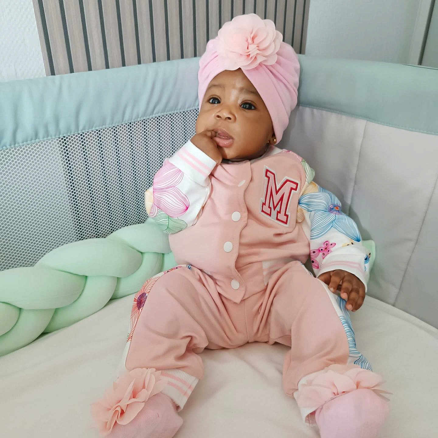 Mariam Sarr a rejoint le concours — aidez-le/la à gagner de superbes lots ! apparel, baby, child, comfortable, cozy, crib, cushion, cute, expression, face, floral_pattern, hand, headwrap, indoors, infant, pink_clothing, portrait, sitting, sock, soft_lighting