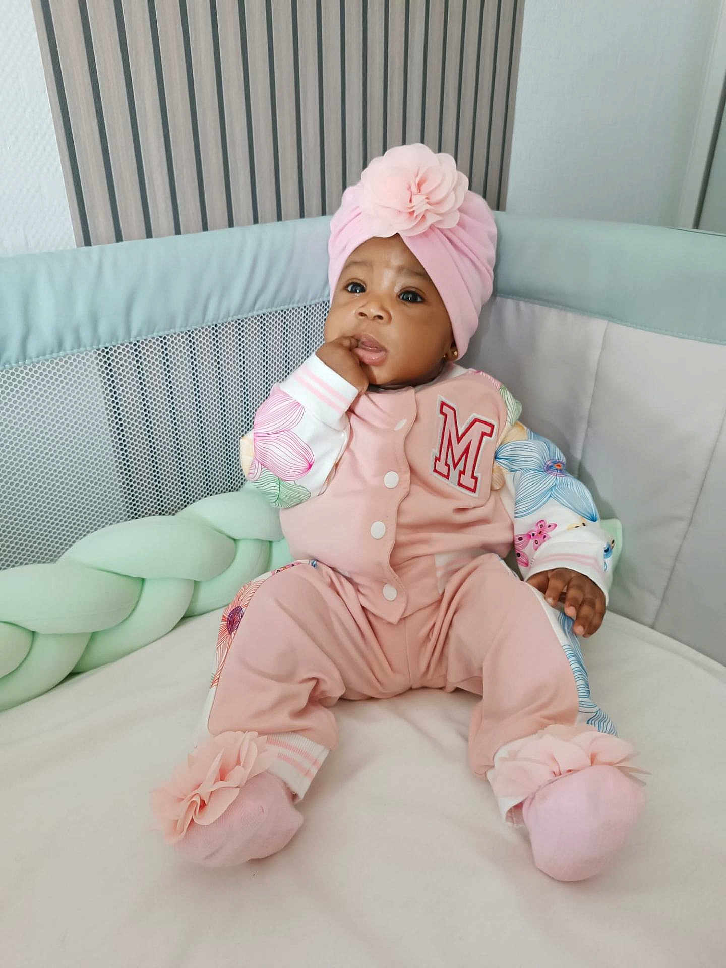 Mariam Sarr a rejoint le concours — aidez-le/la à gagner de superbes lots ! apparel, baby, child, comfortable, cozy, crib, cushion, cute, expression, face, floral_pattern, hand, headwrap, indoors, infant, pink_clothing, portrait, sitting, sock, soft_lighting