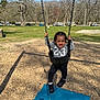 child, toddler, swing, playground, park, outdoor, smiling, happy, black_hair, sneakers, gray_sweatshirt, chains, wood_chips, blue_mat, trees, grass, daylight, person, recreation, fun