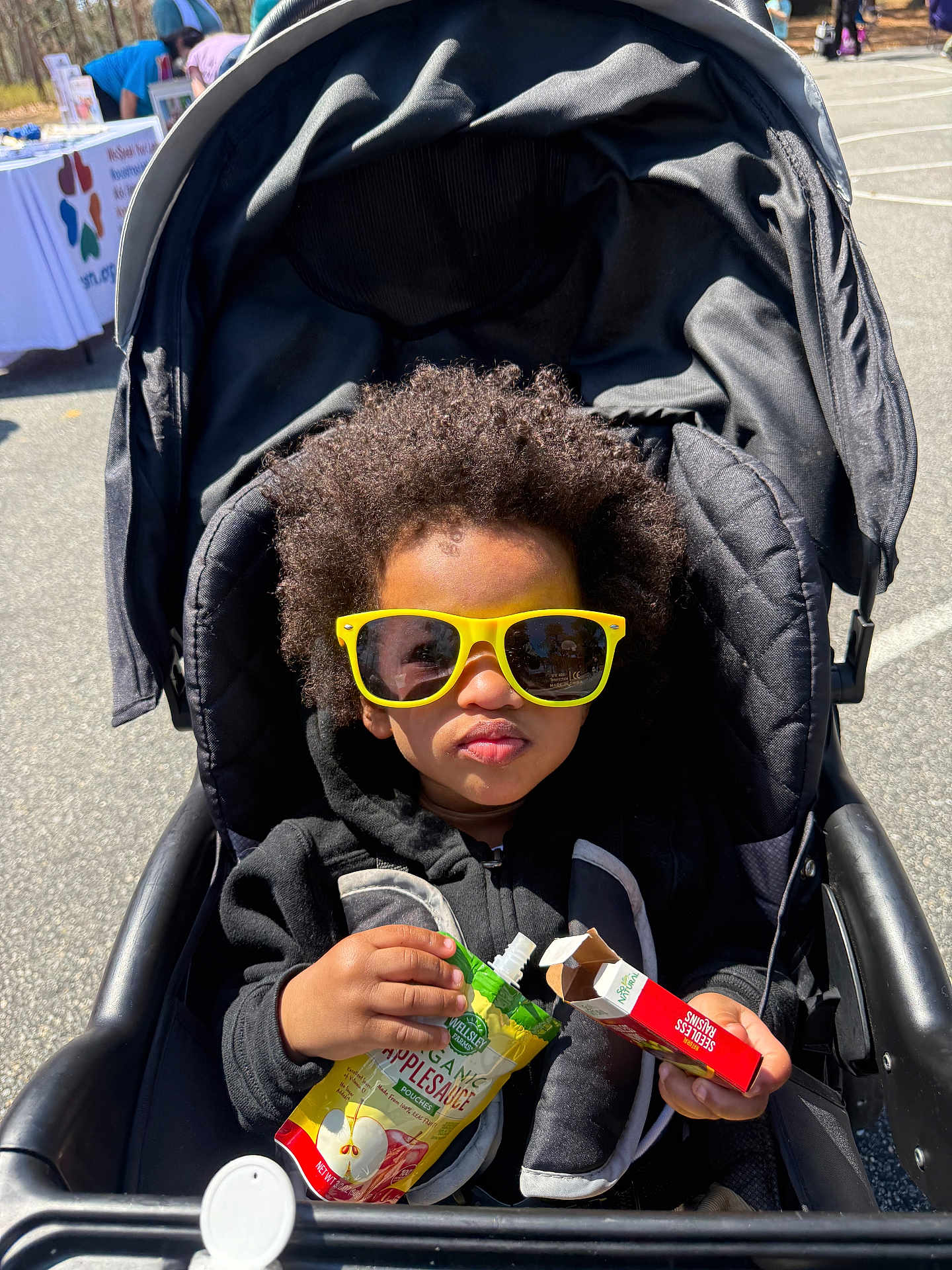 Malakai is registered to the contest to win money with this photo: toddler, child, stroller, sunglasses, yellow_sunglasses, afro_hair, organic_applesauce, seedless_raisins, snack, outdoor, sunny, daytime, person, black_hoodie, baby_food, cute, portrait, food, hand, seat