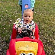 Maddox joined the competition — help win amazing prizes! baby, child, toy_car, outdoor, grass, person, steering_wheel, blanket, colorful_clothing, red, yellow, blue, playtime, park, summer, cute, young_child, shorts, shirt, hands