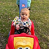 baby, child, toy_car, outdoor, grass, person, steering_wheel, blanket, colorful_clothing, red, yellow, blue, playtime, park, summer, cute, young_child, shorts, shirt, hands