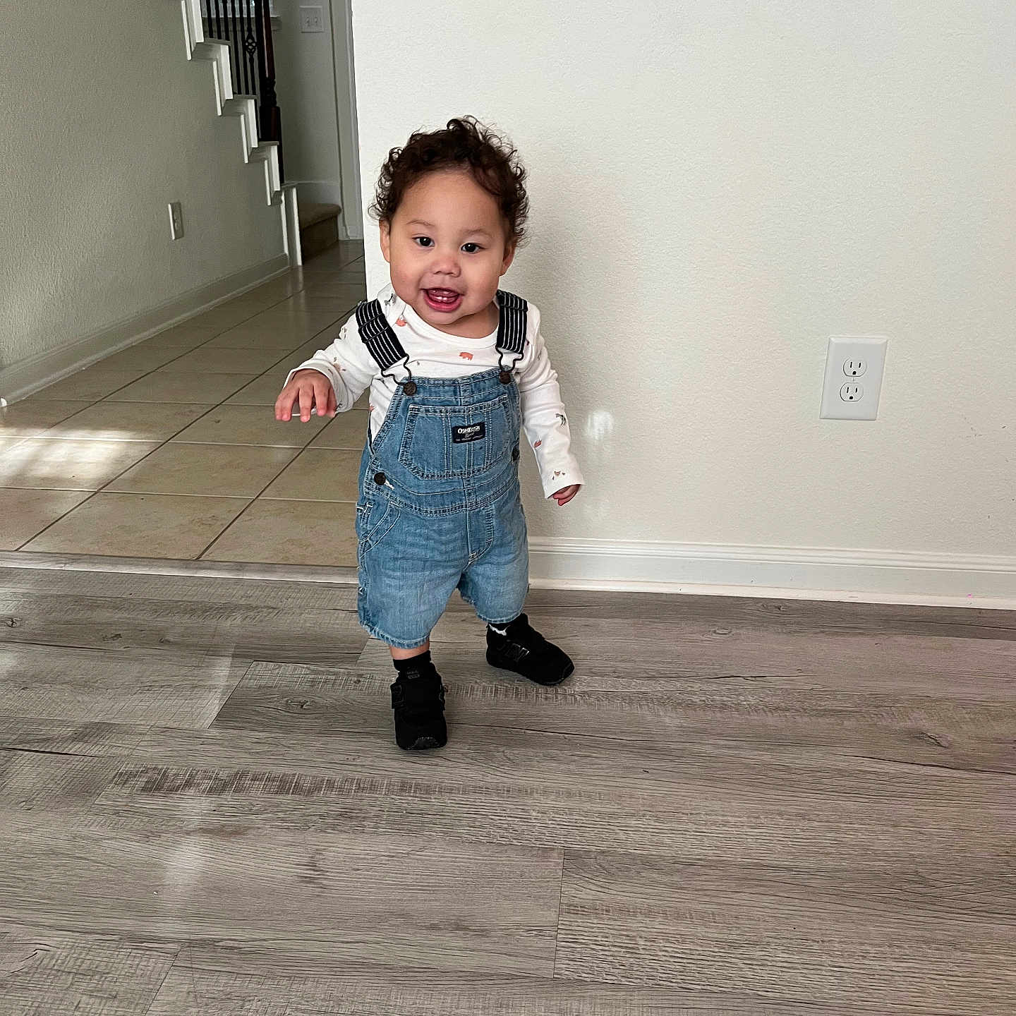 Damián joined the competition — help win amazing prizes! black_shoes, casual_clothing, child, curly_hair, cute, denim_overalls, electrical_outlet, flooring, happy, home, indoor, light, person, portrait, smiling, staircase, standing, toddler, wall, wooden_floor