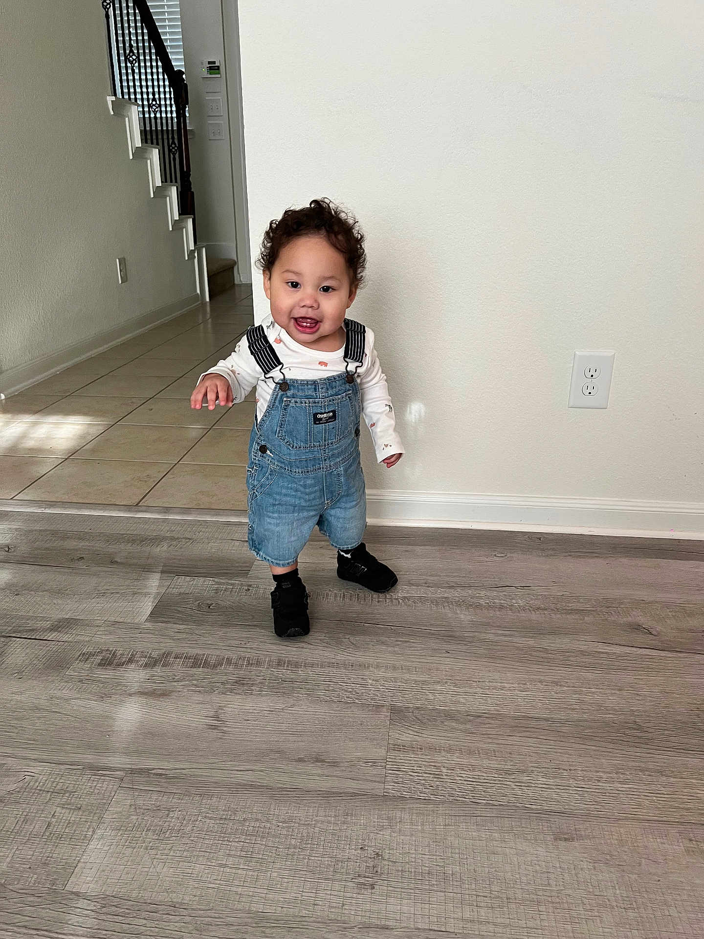 Damián joined the competition — help win amazing prizes! toddler, child, smiling, standing, denim_overalls, black_shoes, curly_hair, indoor, wooden_floor, staircase, wall, electrical_outlet, happy, person, home, casual_clothing, flooring, light, portrait, cute