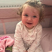 Priscilla is registered to the contest to win money with this photo: toddler, child, smiling, curly_hair, hair_clip, pink_blanket, crib, pajamas, indoors, can, baby, sitting, cute, face, happy, soft_light, cozy, infant, person, portrait
