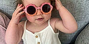 Priscilla joined the competition — help win amazing prizes! toddler, child, baby, sunglasses, pink, white_clothing, romper, bow, hair, couch, gray, fabric, cute, playful, sitting, indoor, person, portrait, smile, fashion