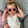toddler, child, baby, sunglasses, pink, white_clothing, romper, bow, hair, couch, gray, fabric, cute, playful, sitting, indoor, person, portrait, smile, fashion