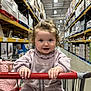 Priscilla is registered to the contest to win money with this photo: toddler, child, shopping_cart, warehouse, store, curly_hair, pink_clothing, sitting, holding, aisle, shelves, products, indoor, portrait, smiling, baby, person, retail, shopping, cute