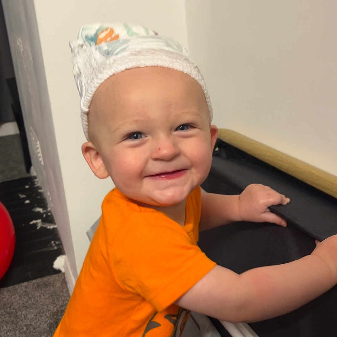 Nolan is registered to the contest to win money with this photo: baby, blue_eyes, carpet, child, cute, diaper, fun, happy, headwear, holding, indoor, infant, orange_clothing, person, playful, small_person, smile, stroller, toddler, wall