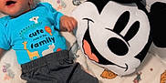 Josue Alexander is registered to the contest to win money with this photo: baby, child, blue_shirt, text_on_clothing, blanket, animal_prints, plush_pillow, mickey_mouse, smiling, lying_down, indoors, cozy, cute, happy, pants, socks, person, infant, toy, colorful