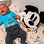 baby, child, blue_shirt, text_on_clothing, blanket, animal_prints, plush_pillow, mickey_mouse, smiling, lying_down, indoors, cozy, cute, happy, pants, socks, person, infant, toy, colorful