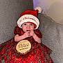baby, infant, child, santa_hat, christmas, holiday, plaid_dress, red_dress, milestone_sign, three_months, wooden_sign, couch, sofa, portrait, indoor, hands_near_mouth, big_eyes, cute, festive, hat_pom_pom