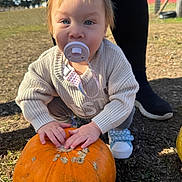 Leilani is registered to the contest to win money with this photo: baby, child, toddler, pacifier, pumpkin, autumn, outdoor, portrait, face, blue_eyes, blonde_hair, sweater, shoes, sitting, hands, garden, grass, playground, adult_legs, daylight