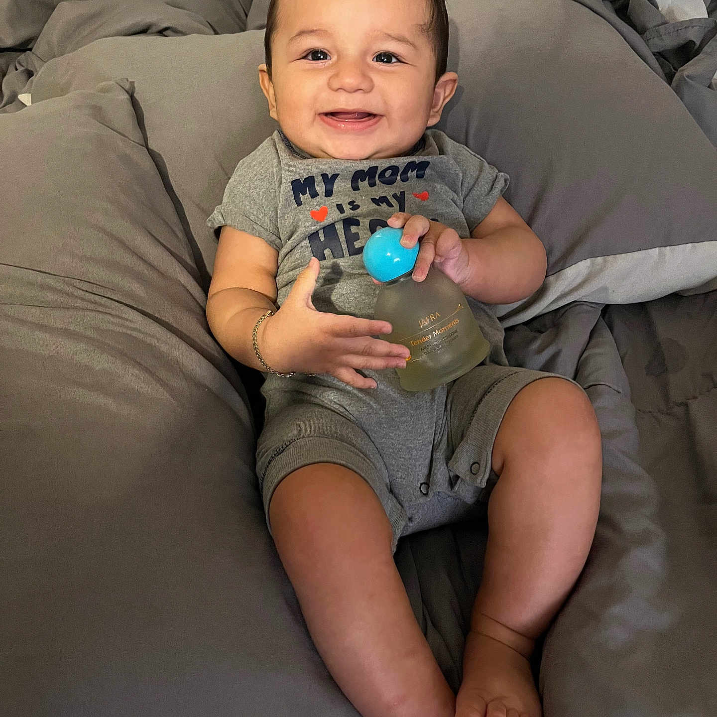 Carlos is registered to the contest to win money with this photo: baby, smiling, infant, onesie, gray, perfume_bottle, blue_cap, cushion, soft_fabric, sitting, happy, child, portrait, indoors, cute, young, skin, person, hand, foot