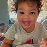 child, toddler, smiling, happy, portrait, indoor, bedroom, bed, blanket, bedding, sitting, curly_hair, dark_hair, gray_shirt, heart_symbol, window, sunlight, backlighting, closeup, photograph