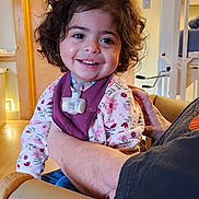 Athena joined the competition — help win amazing prizes! toddler, child, smiling, curly_hair, indoor, person, adult_arm, sweater, floral_pattern, happy, face, seated, holding, home, wooden_door, light_switch, medical_tube, cozy, casual_clothing, expression