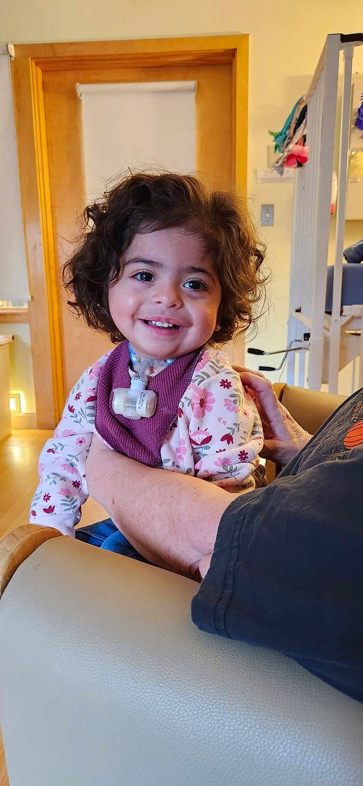 Athena joined the competition — help win amazing prizes! toddler, child, smiling, curly_hair, indoor, person, adult_arm, sweater, floral_pattern, happy, face, seated, holding, home, wooden_door, light_switch, medical_tube, cozy, casual_clothing, expression