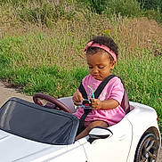 Elyssa a rejoint le concours — aidez-le/la à gagner de superbes lots ! toddler, child, toy_car, pink_clothing, headband, sunglasses, seatbelt, outdoor, greenery, grass, path, curly_hair, concentration, small_vehicle, playtime, summer, nature, side_mirror, parked, casual