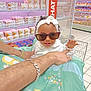 baby, child, sunglasses, headband, adult_hand, grocery_store, shopping_cart, aisle, shelf, products, tattoo, bracelet, white_clothing, floor_tiles, shopping, retail, cute, fashion, smile, indoor
