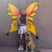 Gemini is registered to the contest to win money with this photo: animal, art, butterfly_wings, casual_clothing, daylight, dog, glasses, happy, leash, mural, outdoor, pet, playful, portrait, sidewalk, smiling, sneakers, tattoos, wall, woman