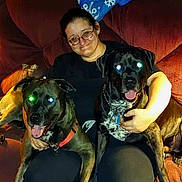 Mischief is registered to the contest to win money with this photo: person, two_dogs, dog, black_dog, brindle_dog, couch, living_room, glasses, collar, tongue_out, smiling, sitting, lap, pet_owner, flash_glare, red_eye, blanket, arm, cozy, indoor