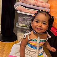 Danna is registered to the contest to win money with this photo: child, toddler, girl, smiling, portrait, indoor, seated, dress, colorful_clothing, flower_hair_clips, braids, medical_tubing, tracheostomy_tube, medical_device, storage_bins, folded_blankets, wooden_floor, fan, home, happy