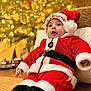 Lucca joined the competition — help win amazing prizes! baby, child, christmas, santa_claus_costume, holiday, festive, hat, red_clothing, white_fur_trim, black_belt, boots, chair, cushion, face, expression, indoor, lights, decorations, tree, celebration