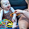 adult, baby, baby_carrier, blue_blanket, child, closeup, colorful_toys, family, happy, indoor, infant, laughing, leopard_print, onesie, orange_ring, playtime, portrait, sitting, smiling, toy