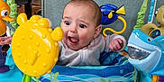 Amelie is registered to the contest to win money with this photo: baby, child, activity_center, toys, yellow_pufferfish, blue_shark_toy, blue_tang_fish_toy, indoor, living_room, couch, playful, surprised_expression, colorful, seat, person, infant, plastic_toys, face, happy, early_childhood