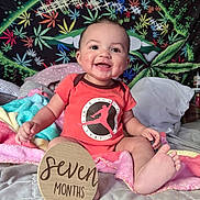 Kaleo is registered to the contest to win money with this photo: baby, bed, blanket, child, colorful, face, foot, hand, happy, indoors, onesie, orange_clothing, pillow, plant_pattern, portrait, seven_months, sitting, smiling, tapestry, wooden_sign