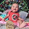 baby, child, smiling, onesie, orange_clothing, blanket, bed, pillow, wooden_sign, seven_months, face, happy, sitting, indoors, tapestry, colorful, plant_pattern, foot, hand, portrait