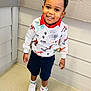 toddler, child, smiling, braided_hair, sweatshirt, dinosaurs, shorts, sneakers, socks, indoor, wall, corner, floor, happy, person, standing, portrait, casual_clothing, cute, playful