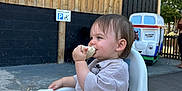 Teddy is registered to the contest to win money with this photo: toddler, child, high_chair, sandwich, eating, outdoor, wooden_wall, sign, play_area, ride, person, happy, casual, daylight, smile, short_hair, clothing, seat, background, fun
