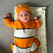 Albie is registered to the contest to win money with this photo: baby, child, costume, orange, clownfish, hat, tongue_out, lying_down, carpet, patterned_mat, cute, playful, face, eyes, hands, infant, clothing, indoor, person, smile