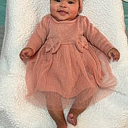 Amara is registered to the contest to win money with this photo: baby, child, smiling, pink_dress, headband, bow, blanket, soft_texture, feet, happy, cute, infant, portrait, indoors, clothing, toddler, lying_down, person, fashion, cozy