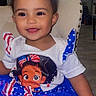 Saphira is registered to the contest to win money with this photo: baby, child, infant, smiling, tongue_out, portrait, sitting, indoor, chair, white_chair, dress, stars_and_stripes, american_flag_pattern, cartoon_character, colorful_clothing, patriotic, curly_hair, brown_eyes, flash_photography, home