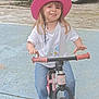 child, pink_hat, white_tshirt, balance_bike, outdoor, pavement, car, greenery, happy, smiling, sandals, blue_jeans, toy_bike, playtime, summer, kid, casual_clothing, portrait, daylight, young_child