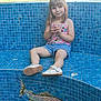 child, girl, smile, sitting, blue_tiles, mosaic, fish_design, outdoor, grass, tree, fence, shoes, shorts, tank_top, person, daylight, casual_clothing, relaxed, portrait, background