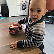 Raphaël a rejoint le concours — aidez-le/la à gagner de superbes lots ! toddler, child, toy, fire_truck, table, striped_sweater, indoor, floor, cabinet, panda_decor, clothing, person, curious, play, wooden_table, shoes, home, furniture, expression, room