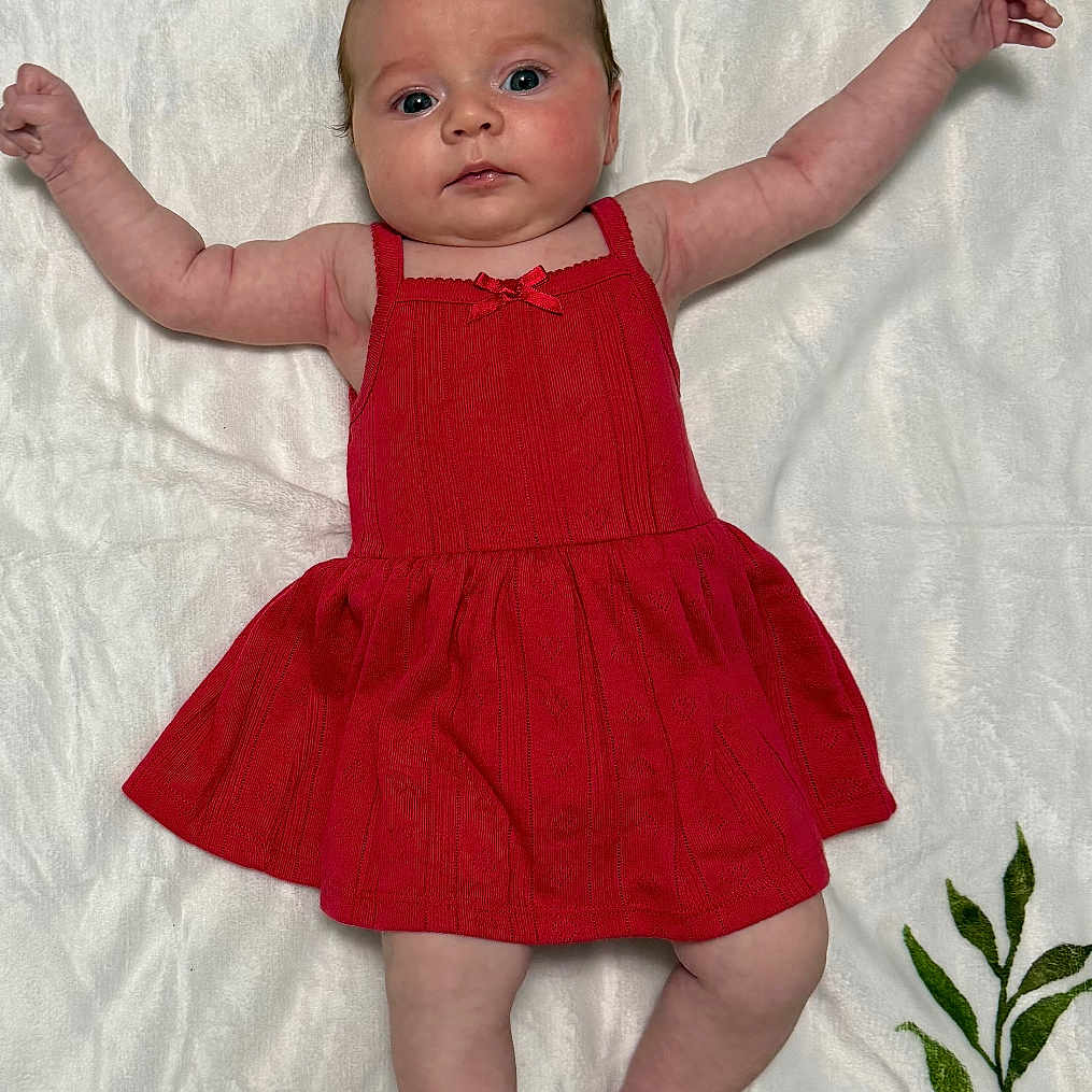 Ava is registered to the contest to win money with this photo: baby, infant, red_dress, floral_blanket, lying_down, arms_outstretched, cute, child, portrait, indoors, skin, face, expression, feet, head, person, newborn, fabric, pattern, colorful
