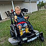building, child, daylight, grass, grill, hands, happy, kid_trax, lawn, outdoor, overalls, patio_chairs, person, play, rain_boots, seatbelt, smiling, toddler, toy_vehicle, yellow_boots