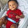 baby, infant, child, smiling, red_clothing, onesie, socks, baby_seat, cushion, indoor, face, hand, legs, curly_hair, toddler, comfort, seatbelt, floor, wooden_floor, portrait