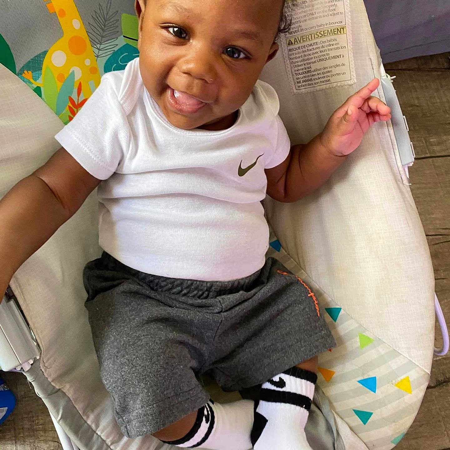 Markylan joined the competition — help win amazing prizes! baby, child, smiling, sitting, clothing, white_shirt, gray_shorts, socks, nike_logo, baby_seat, giraffe_pattern, indoor, flooring, happy, cute, portrait, infant, playful, person, young