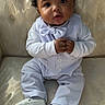 Amari is registered to the contest to win money with this photo: baby, child, infant, suit, bow_tie, white_clothing, shoes, curly_hair, couch, upholstery, patterned_fabric, seated, hands_clasped, looking_at_camera, indoors, portrait, cute, young_child, formal_wear, small_shoes