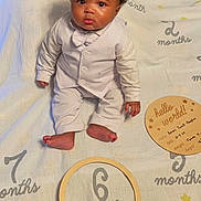 Amari is registered to the contest to win money with this photo: baby, infant, child, milestone, white_suit, blanket, floor, wood_floor, sitting, portrait, indoors, cute, face, curly_hair, expression, six_months, clothing, person, young_child, milestone_marker