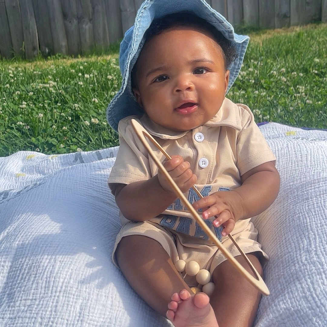 Amari is registered to the contest to win money with this photo: baby, child, outdoor, grass, blanket, hat, toy, cute, smiling, sunlight, portrait, infant, happy, sitting, wooden_toy, feet, clothing, nature, fence, daylight