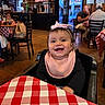 baby, bow, casual_clothing, chairs, checkered, child, dining, face, happy, headband, high_chair, indoor, people, pink_bib, portrait, red_and_white, restaurant, smiling, tablecloth, wooden_wall