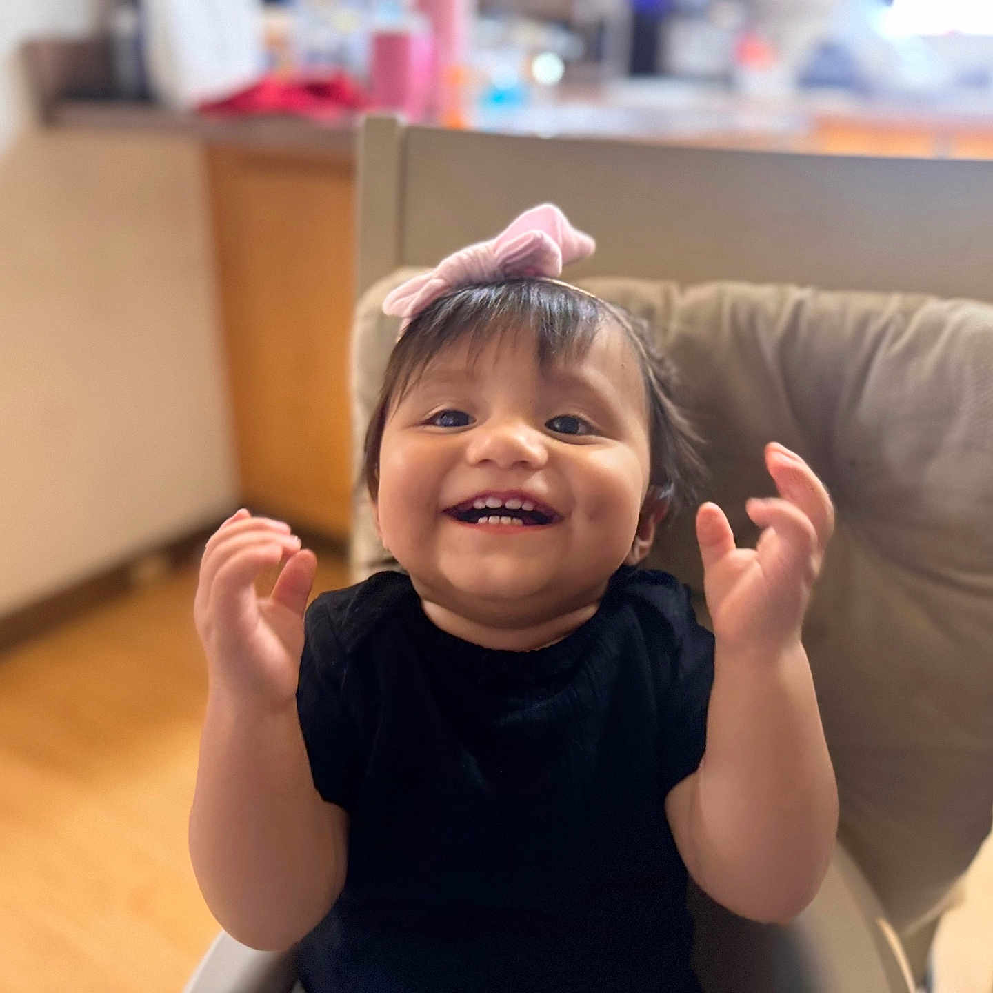 Mykenzie is registered to the contest to win money with this photo: toddler, child, smiling, happy, high_chair, bow, black_shirt, indoors, kitchen, cushion, hands, face, baby, cute, young_child, wooden_floor, furniture, person, portrait, celebration
